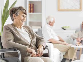 Bild einer fröhlichen Seniorin im Rollstuhl, im Hintergrund unterhalten sich zwei ältere Herren